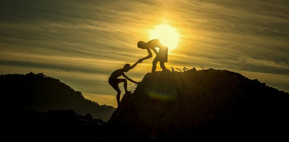 Two people helping each other reach the top of a mountain.